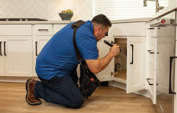 kitchen-inspection buyer home inspection performing a detailed kitchen plumbing evaluation, kneeling to use a high-powered flashlight to check for leaks and material defects under a white shaker-style sink cabinet.