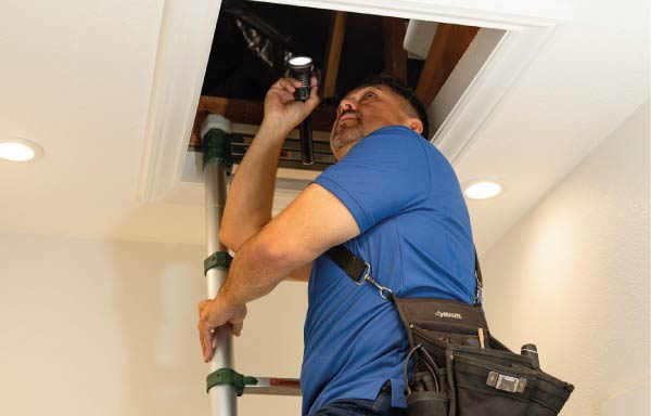 Certified property inspector Oscar Fregoso using a telescopic ladder and flashlight to conduct a thorough attic and roofing structural inspection, identifying safety concerns and insulation conditions.
